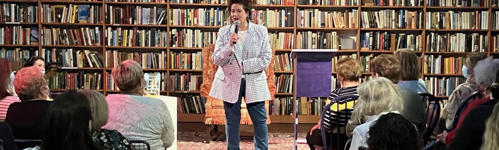 photo of woman with microphone and some audience members against background of bookshelves