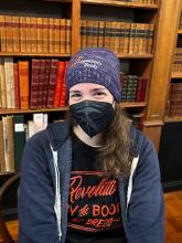 Woman wearing a mask and a purple beanie with LOGANBERRY BOOKS text poses in front of a bookshelf